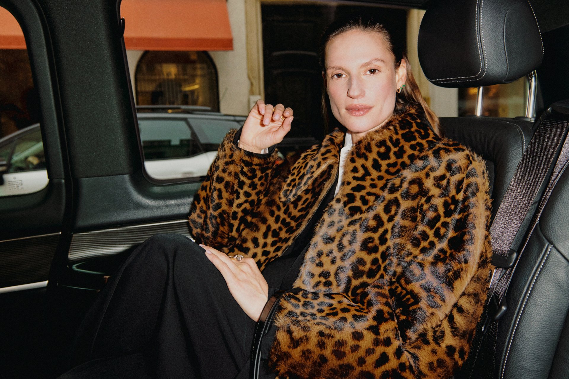 Una persona con un abrigo de piel de leopardo se sienta en un coche, descansando casualmente la mano en su barbilla, con una vista de la ciudad afuera.