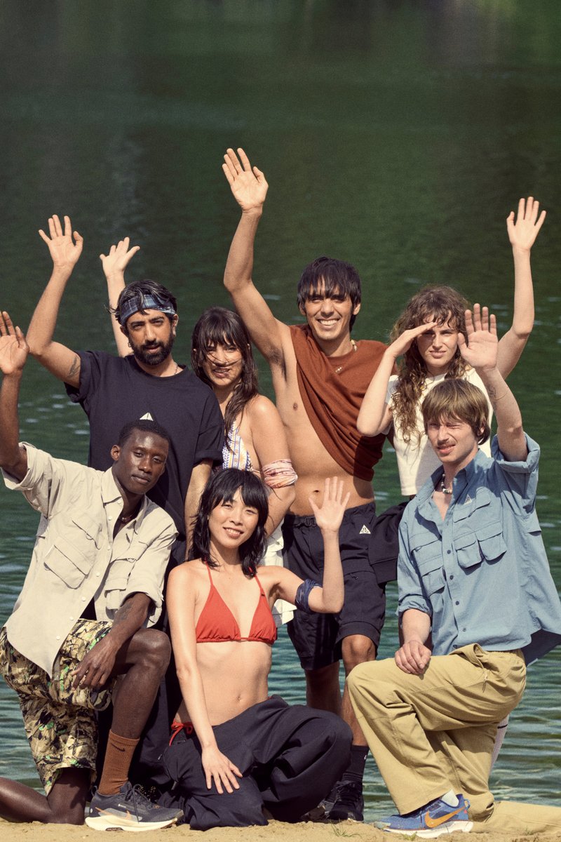 Un gruppo diversificato di otto persone si trova accanto a un lago, sorridendo e salutando. Indossano abbigliamento estivo casual, con colori e stili vivaci.