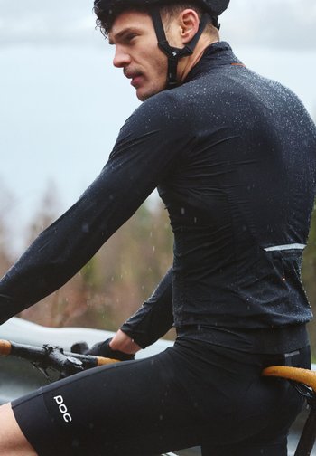 Syklist iført svart regnjakke og hjelm sykler i vått vær, holder fast i bukkestyret med regndråper synlige på klærne.