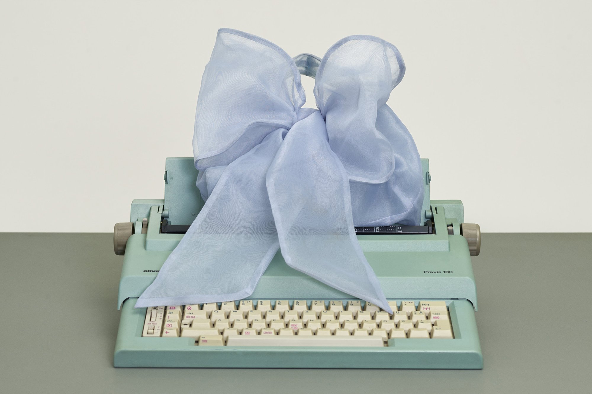 Escribir a máquina de color verde menta con teclas crema, rematada con un gran lazo de gasa azul claro, reposando sobre una superficie gris frente a un fondo neutro.