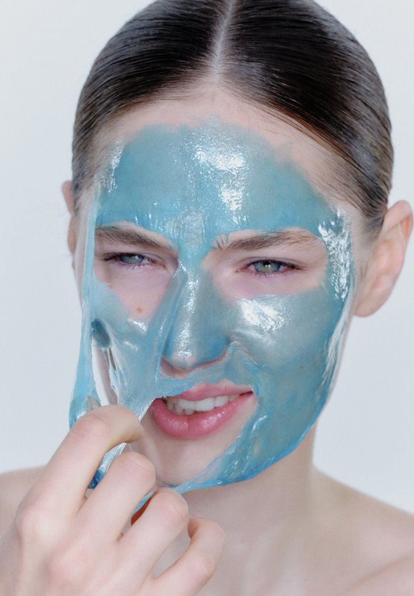 Blue gel face mask being peeled off skin, showcasing a shiny, glossy texture. The model has light skin and is smiling slightly.