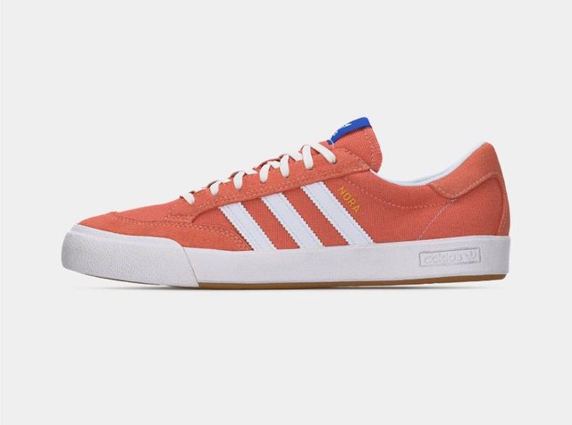 Sneaker orange Adidas à profil bas avec trois bandes blanches, lacets blancs, étiquette bleue sur la languette et semelle en caoutchouc blanche.