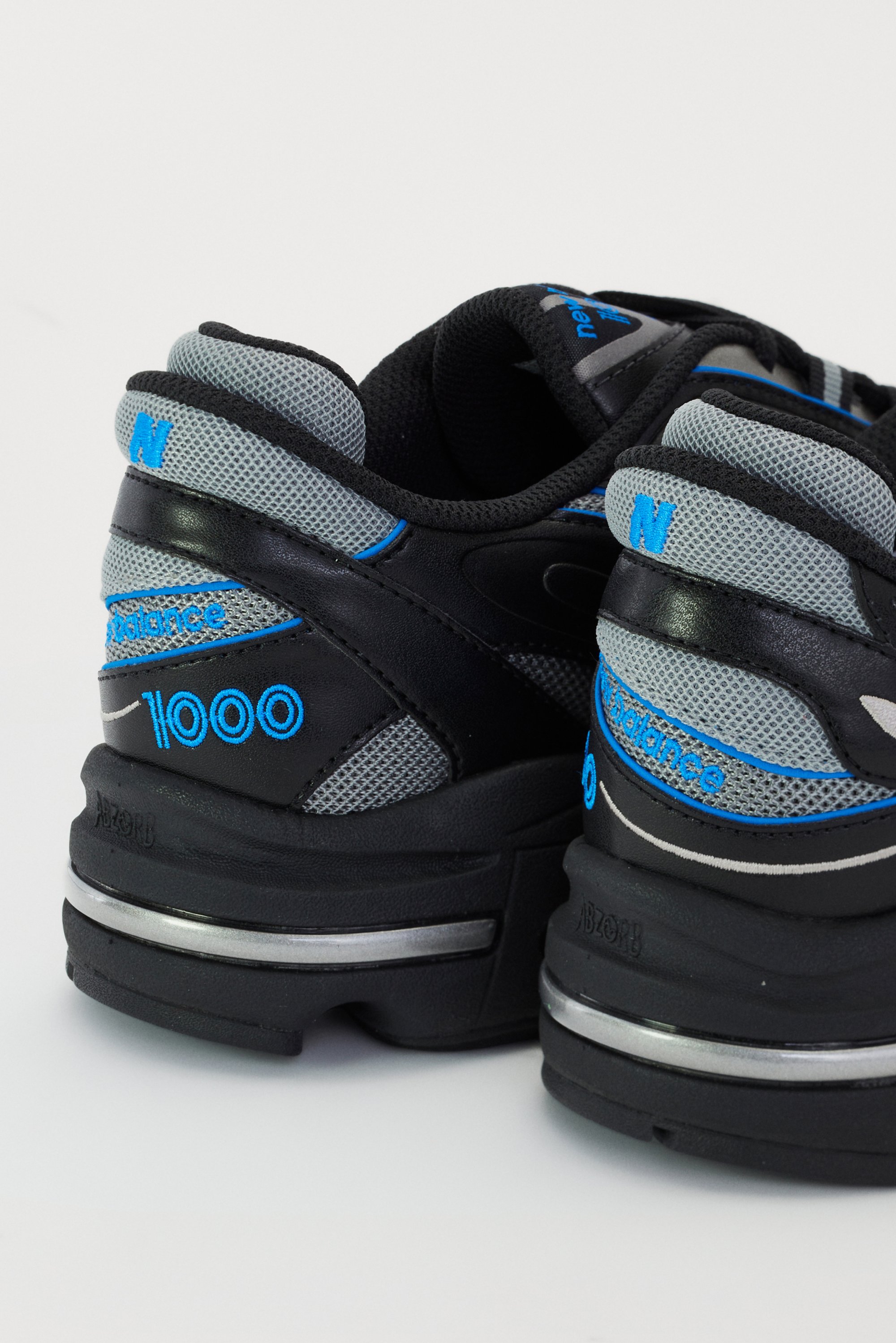 Scarpe da ginnastica nere e grigie con dettagli blu, dotate di un logo "1000" ben visibile e design in rete testurizzata, presentate su uno sfondo bianco.