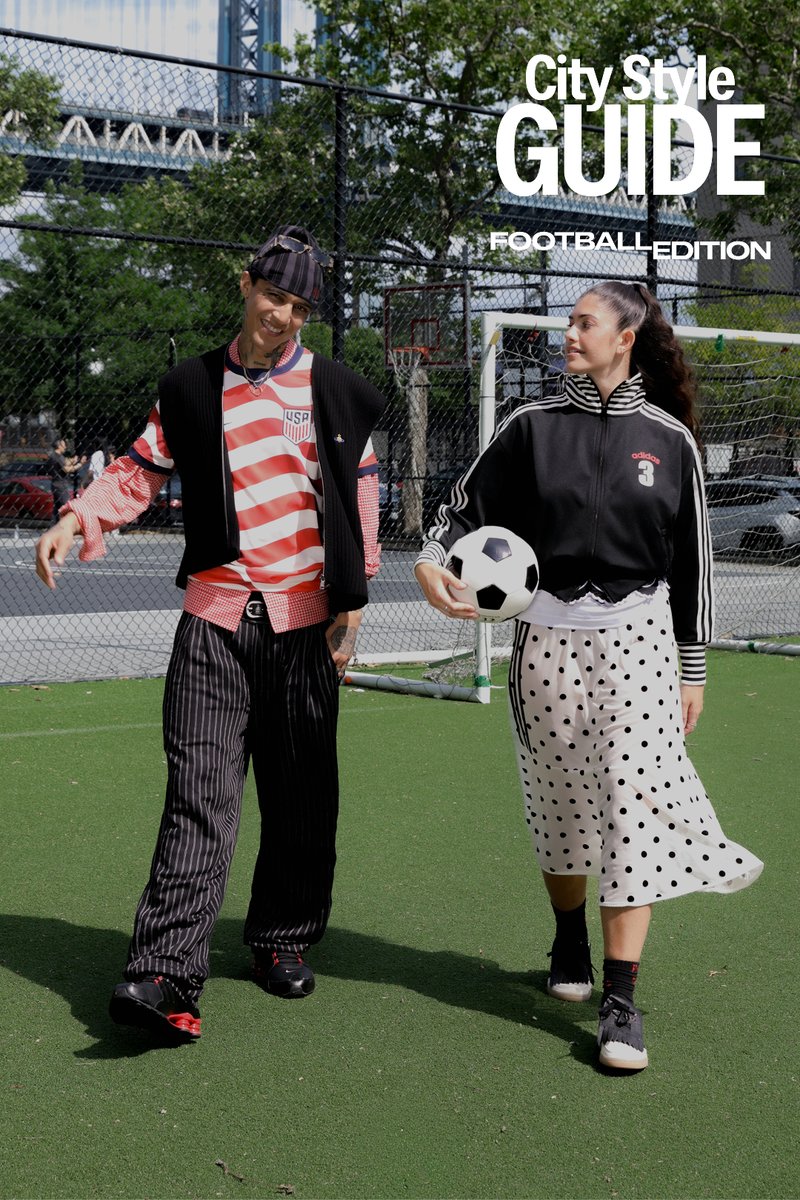 Zwei Personen gehen über ein Fußballfeld, einer hält einen Fußball, beide tragen sportliche und legere Kleidung, im Hintergrund sind eine Stadtsilhouette und ein Tor zu sehen.