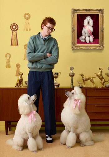 Mann mit Brille, der zwischen zwei weißen Pudeln mit rosa Schleifen steht, Trophäen und Bänder auf einem Schrank, Porträt eines gekrönten Pudels mit Schärpe an der Wand.