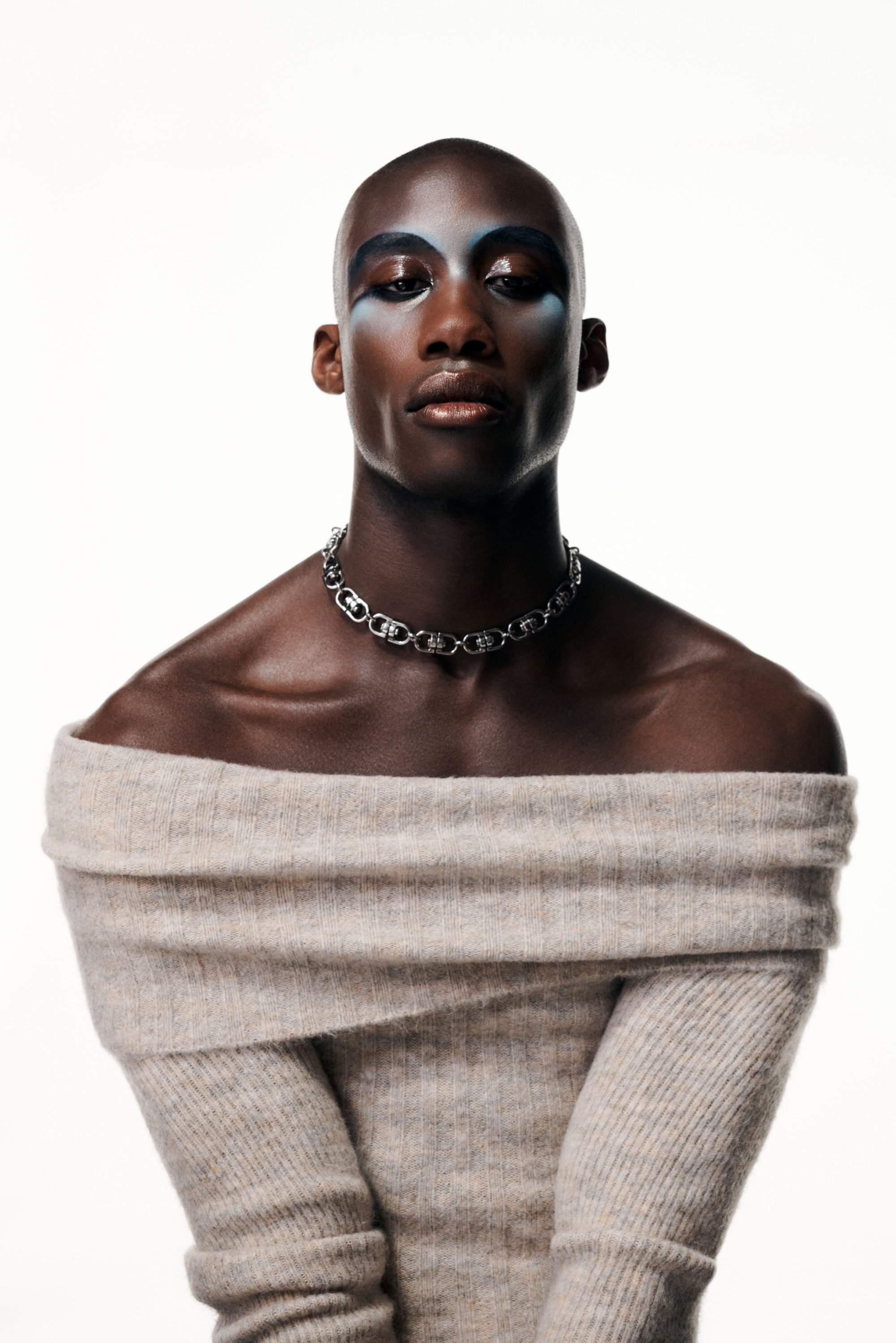 Una persona con la cabeza rapada y un atrevido maquillaje azul en los ojos posa con un suéter, adornado con un collar de cadena de plata gruesa, frente a un fondo blanco.