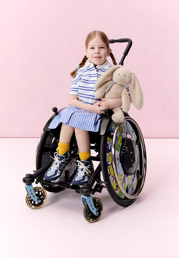 Junges Mädchen mit geflochtenem Haar in gestreiftem blauem Kleid sitzt in einem dekorierten Rollstuhl und hält ein beiges Plüschtierhäschen vor einem rosa Hintergrund.