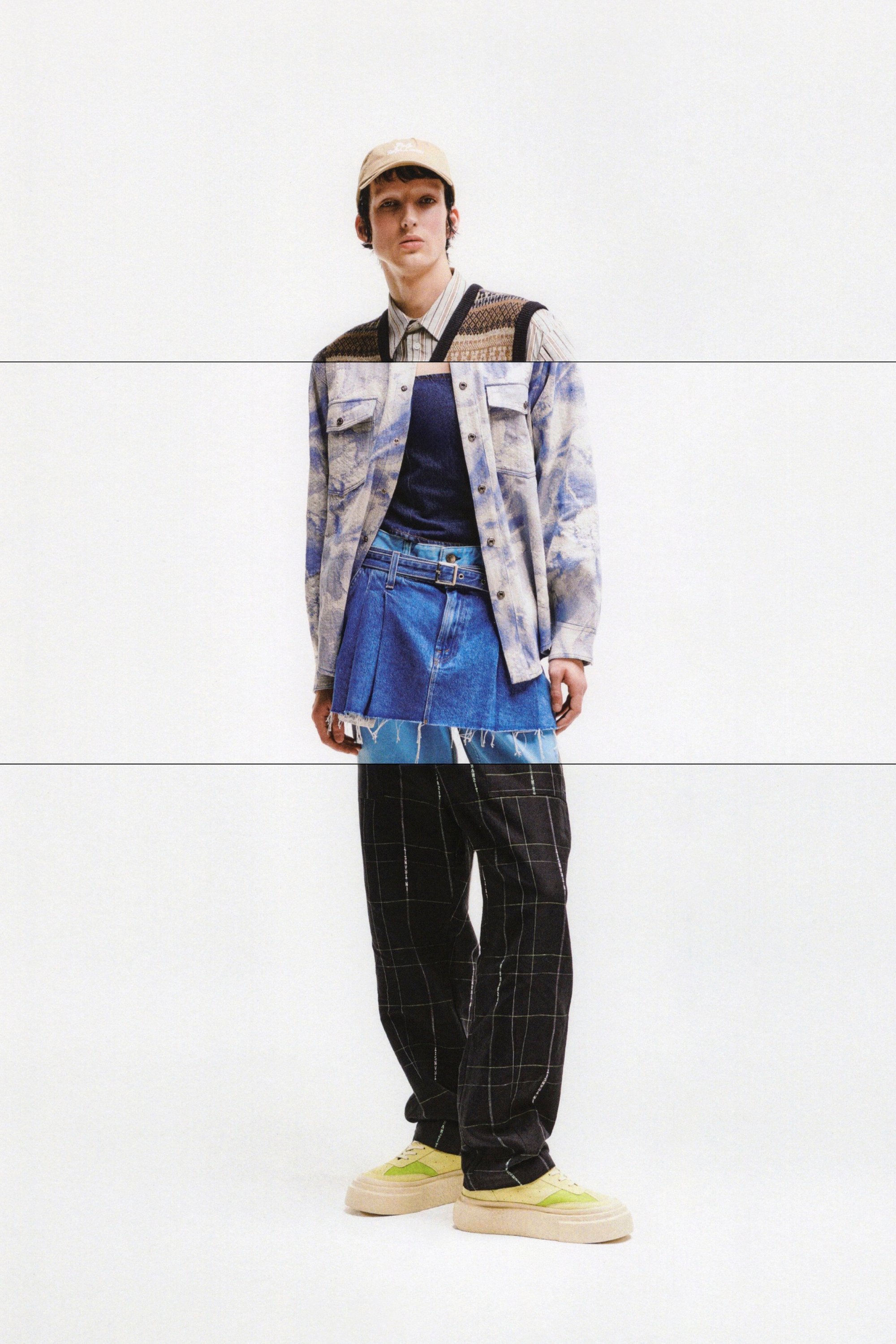 A person stands against a white background, wearing a blue patterned jacket, layered denim skirt, black plaid pants, and bright sneakers.
