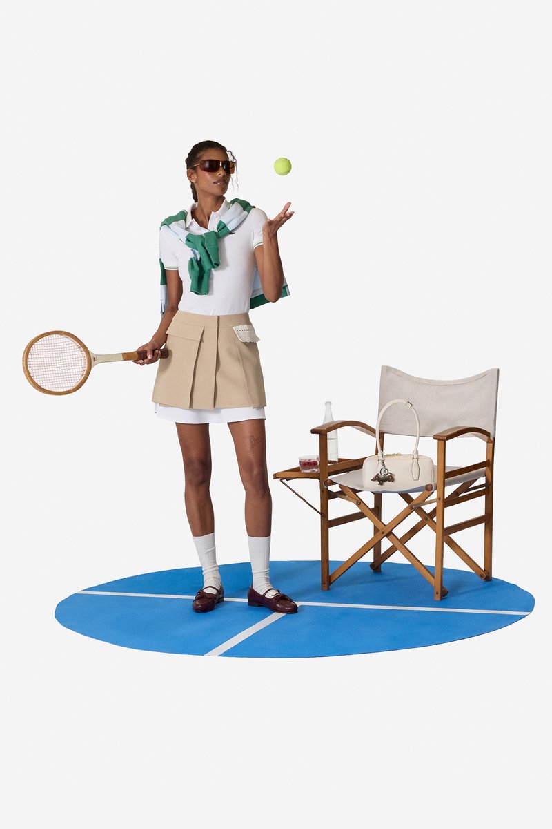 Frau in Tennisoutfit wirft einen Tennisball, hält einen Holz-Tennisschläger und steht neben einem Regiestuhl mit Handtasche und Getränken.