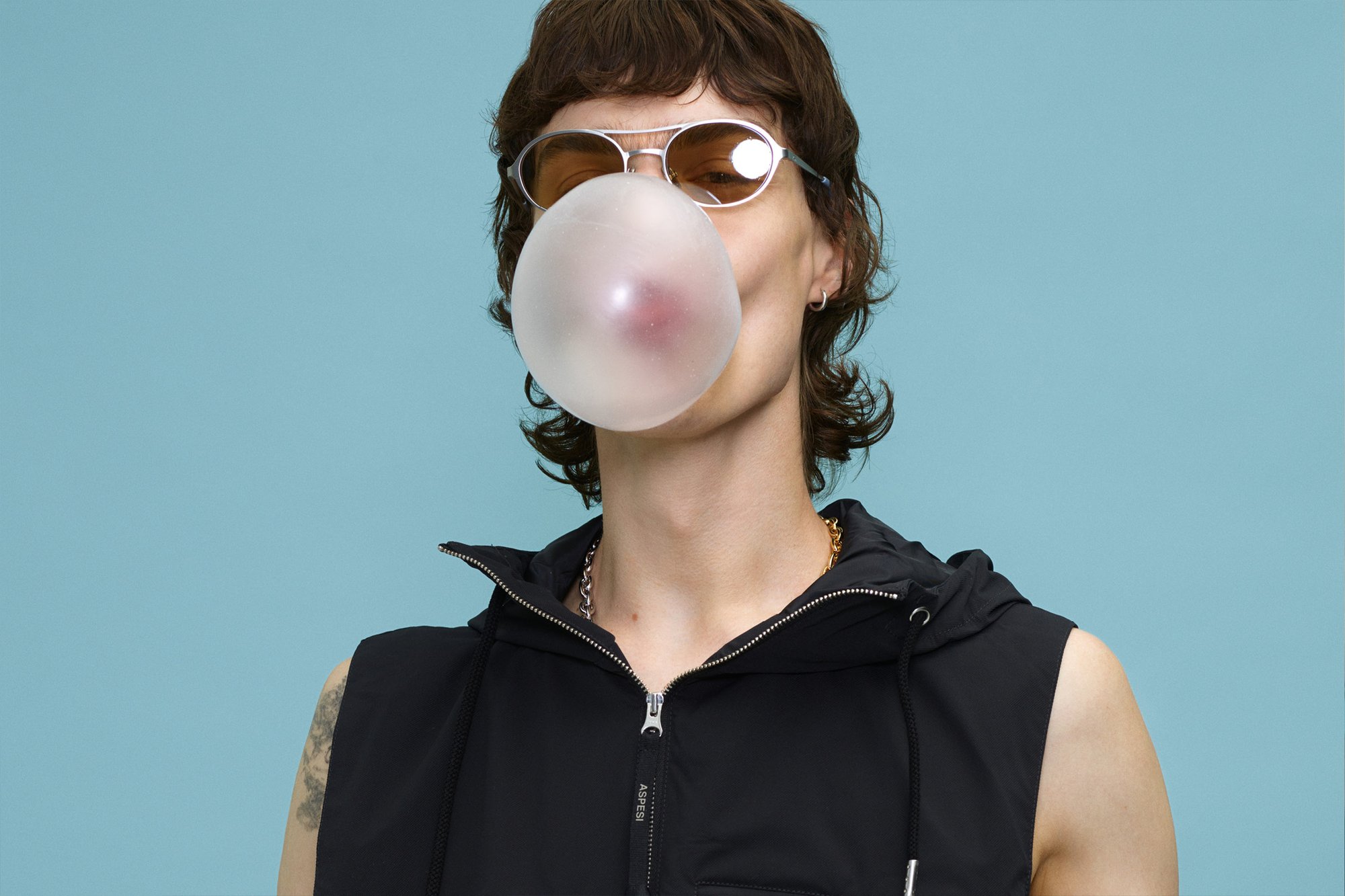Chaqueta sin mangas negra con cremallera en la parte delantera y capucha, gafas de sol plateadas, y una gran burbuja de chicle translúcida contra un fondo azul.
