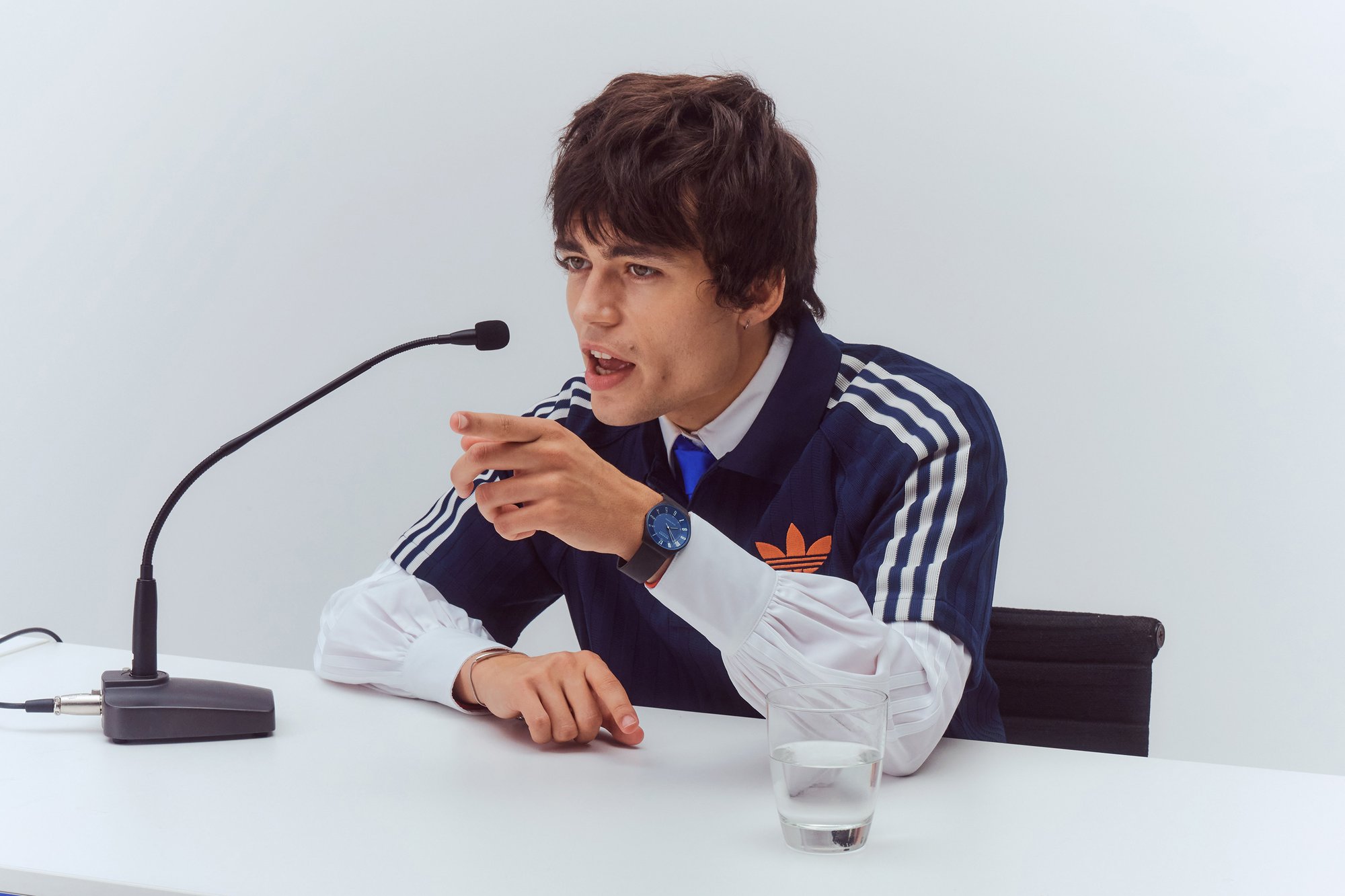 A young man in a dark tracksuit gestures animatedly while speaking into a microphone, with a glass of water on the table.