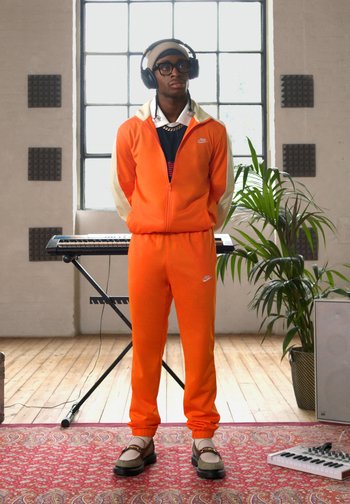Survêtement orange Nike avec des accents blancs, chemise noire en dessous et écouteurs. Comprend des baskets avec une tige grise et une semelle noire épaisse.