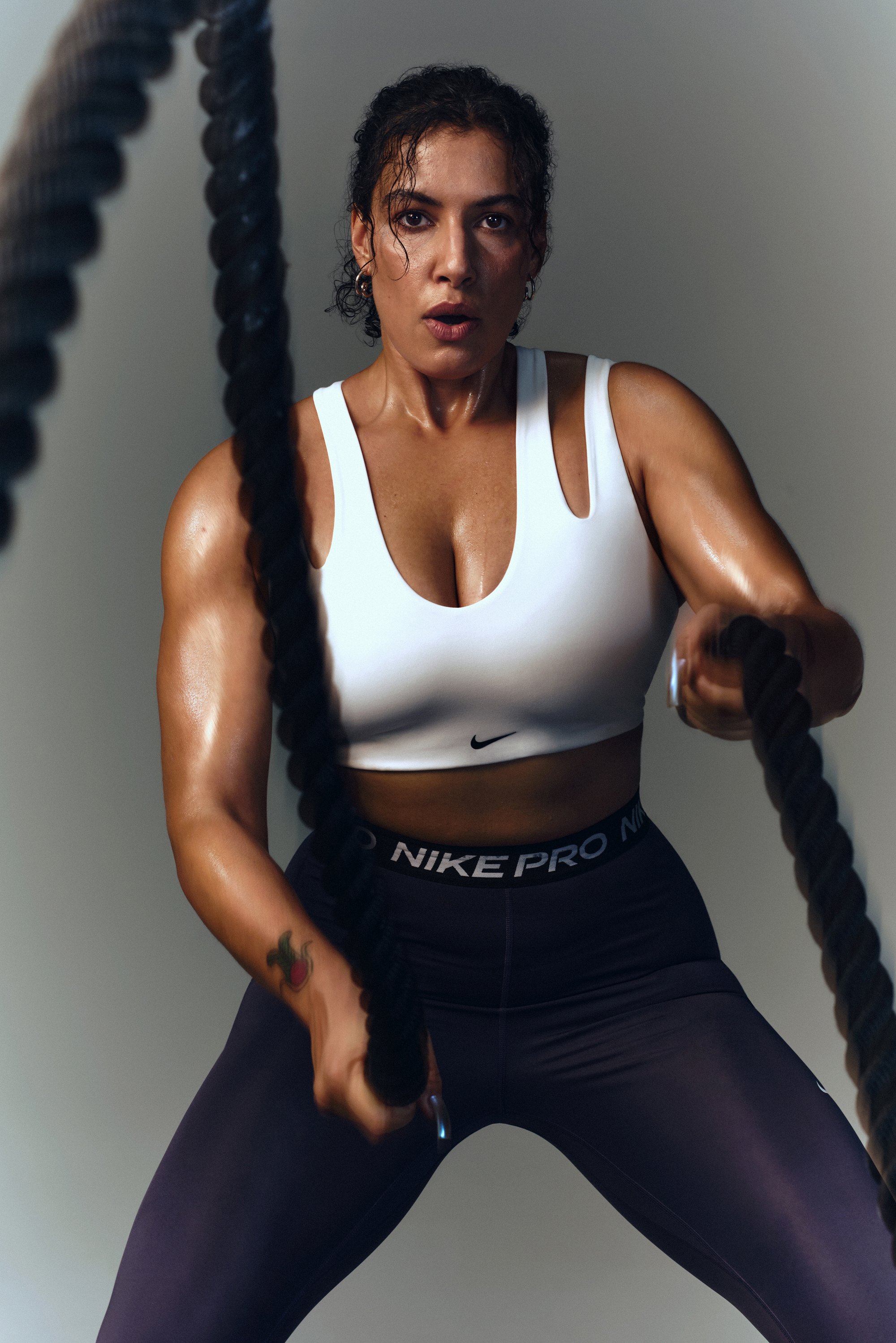 A muscular woman with wet skin and curly hair vigorously swings battle ropes while wearing a white sports bra and black leggings.
