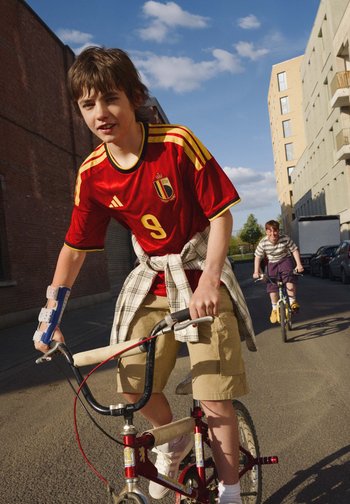 Jongen met onderarmbrace rijdt fiets en draagt een rood Belgisch voetbalshirt, met een andere jongen die achter hem fietst op een zonnige stedelijke straat.