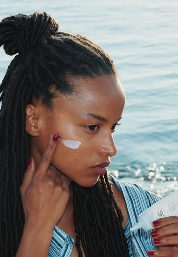 Woman with long braids applying white cream on her cheek near a body of water, holding a tube of skincare product.