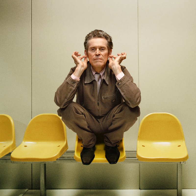 A man in brown clothing squats on a yellow chair, holding his ears, with an expression of concentration, against a minimalistic background.