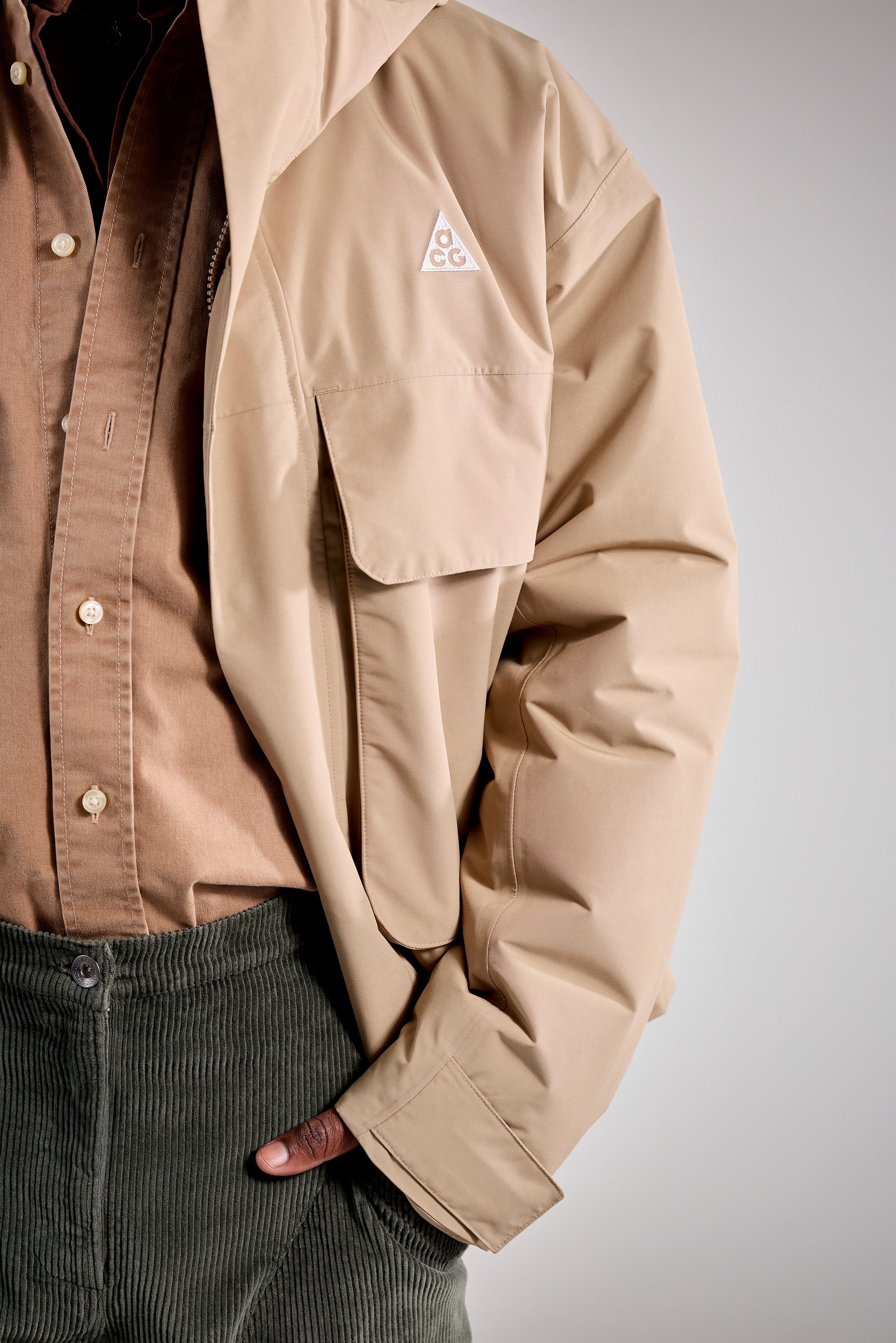 Person wearing a tan jacket with a logo, a brown shirt, and dark green corduroy pants. Hand is casually placed in the jacket pocket.