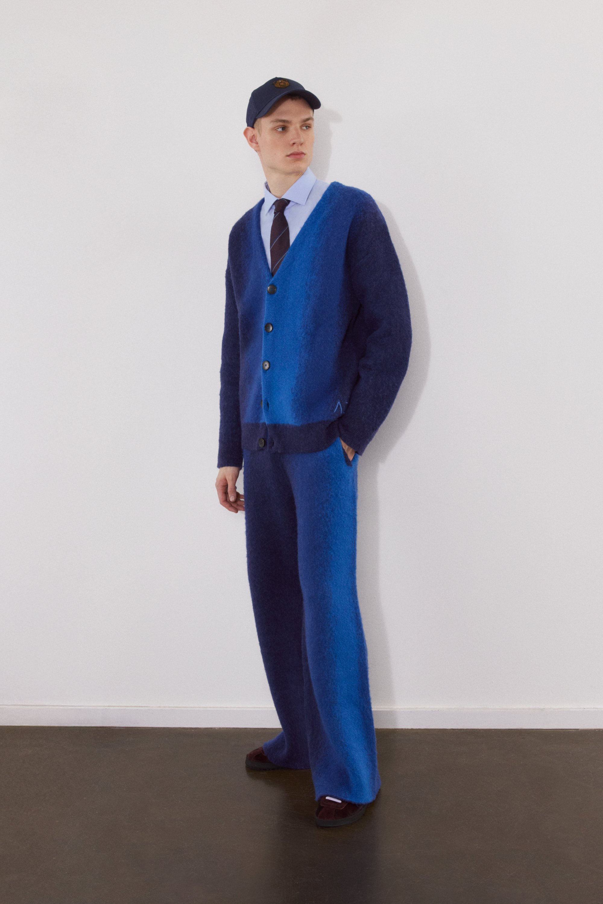 A young man stands against a white wall, wearing a blue gradient cardigan, tailored pants, a light blue shirt, and a navy cap.