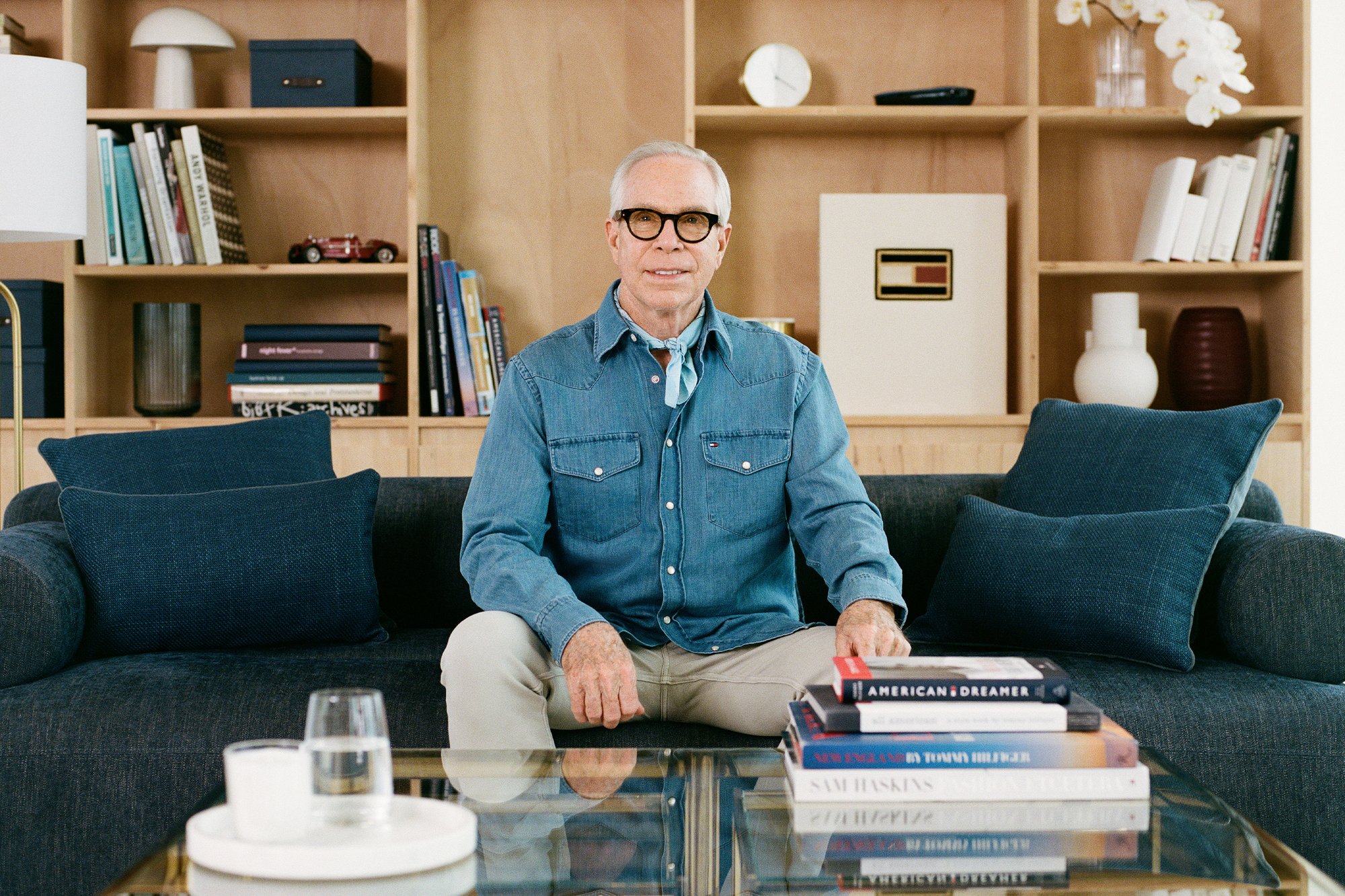 Denim overhemdbloes, lichtblauw. Beige broek. Donkergrijze bank met twee marineblauwe kussens. Glazen salontafel met gestapelde boeken en een glas.