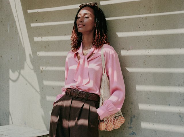 Femme les yeux fermés appuyée contre un mur, portant un chemisier rose, un pantalon marron, et portant un sac en filet avec des oranges au soleil.