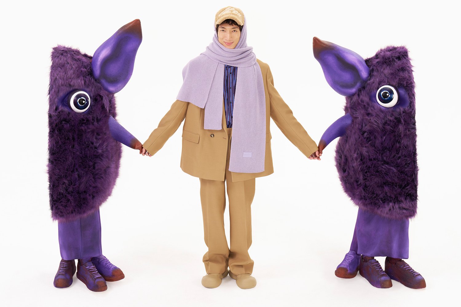 A person in a tan suit and purple scarf holds hands with two large, furry, purple characters with big eyes, set against a white background.