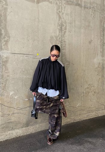 Une femme portant des lunettes de soleil pose devant un mur en béton. Elle porte une veste noire à rayures et un pantalon camouflage, et a deux sacs à main avec elle.