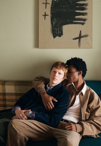 Twee jonge mannen zitten dicht bij elkaar op een sofa, waarbij de één zijn arm om de schouder van de ander slaat. Achter hen hangt een doek met zwarte abstracte kruisen.
