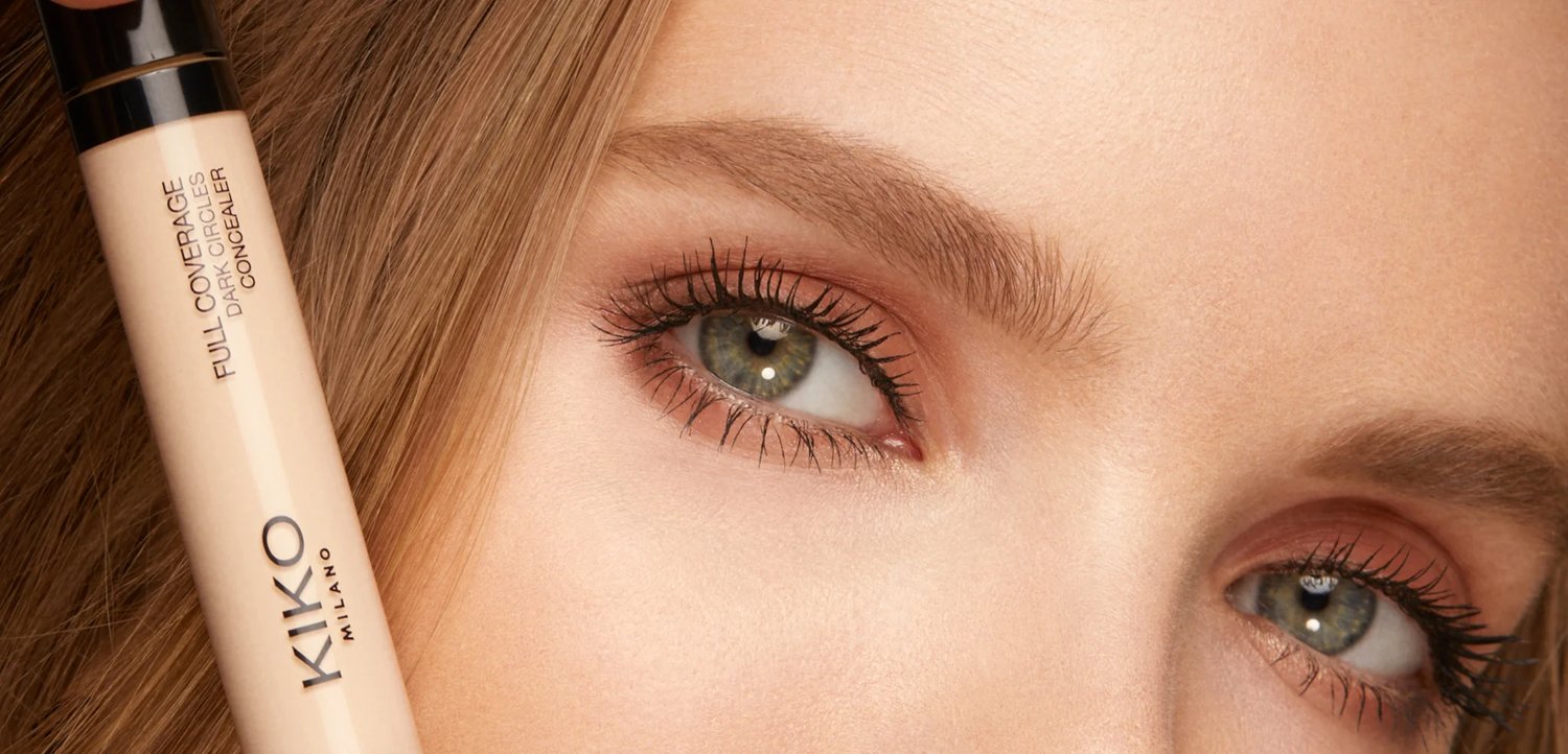 Close-up of green eyes with long lashes and light makeup, next to a beige Kiko Milano Full Coverage concealer stick.