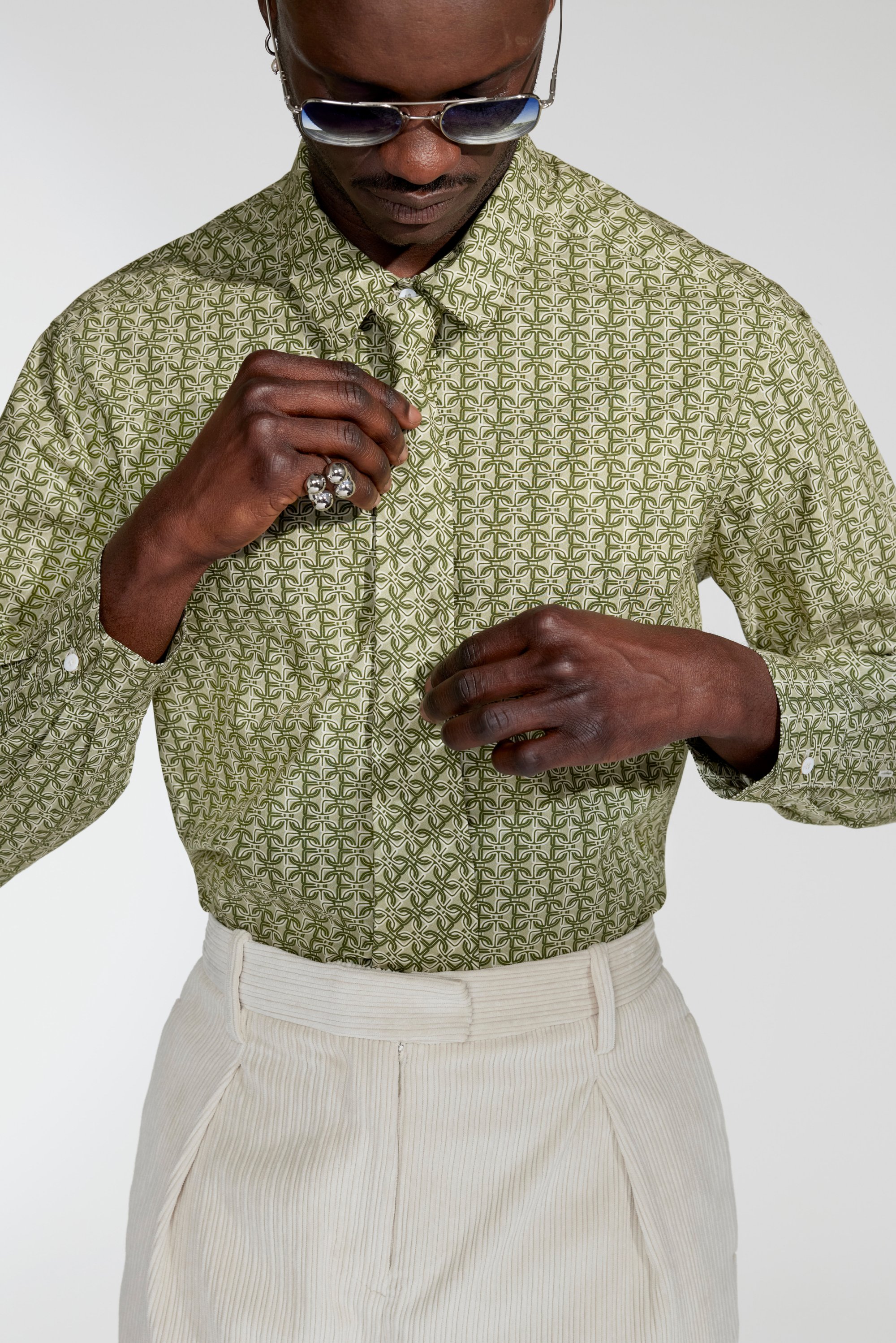 A person adjusts their patterned green shirt, wearing sunglasses and silver rings, paired with pleated beige trousers against a neutral background.