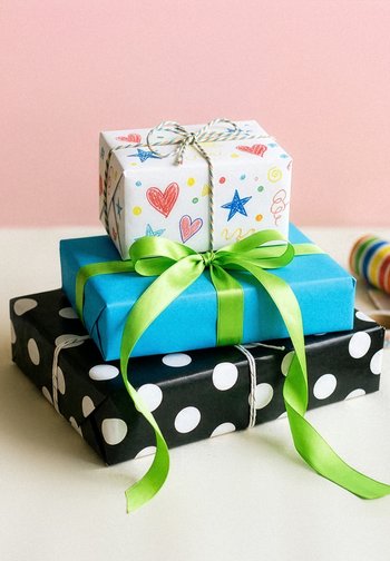 Tri zložene darilne škatle: zgornja bela s pisanimi srcami in zvezdami, srednja modra z zelenim trakom, spodnja črna s belimi pikami.