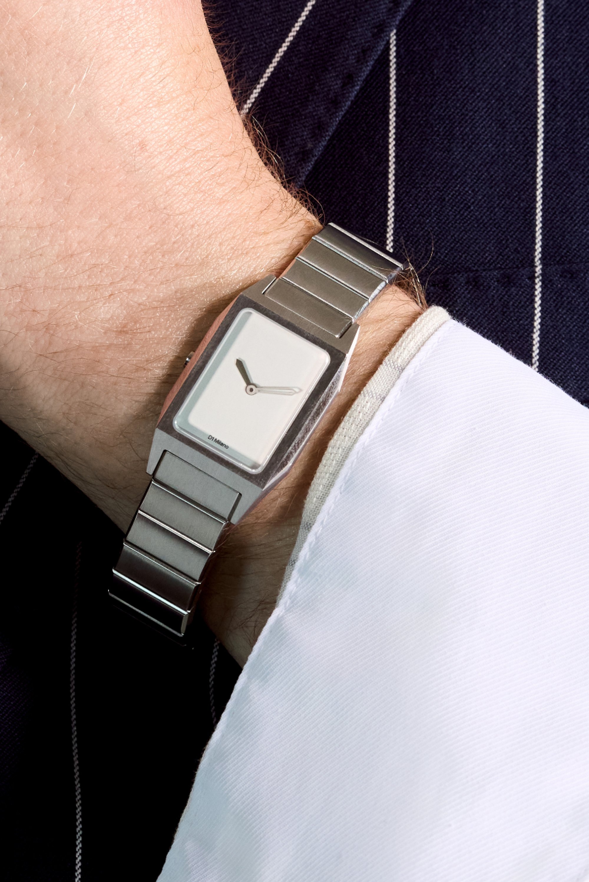 Reloj rectangular plateado con una esfera blanca y manecillas delgadas, con una correa de metal. Lleva puesto en una muñeca al lado de una camisa blanca.