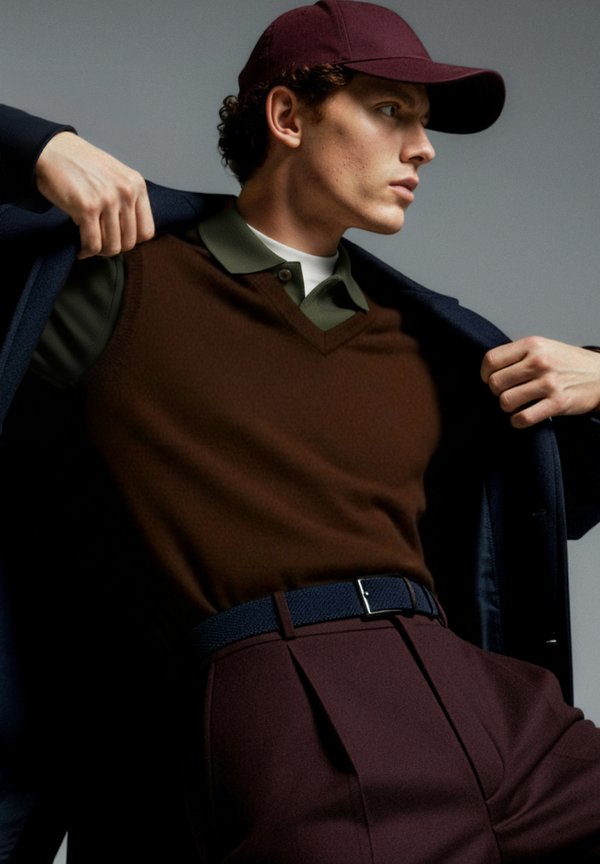 Brun stickad väst över en grön pikétröja, kombinerad med skräddarsydda vinröda byxor och en marinblå kappa. En vinröd keps kompletterar looken.