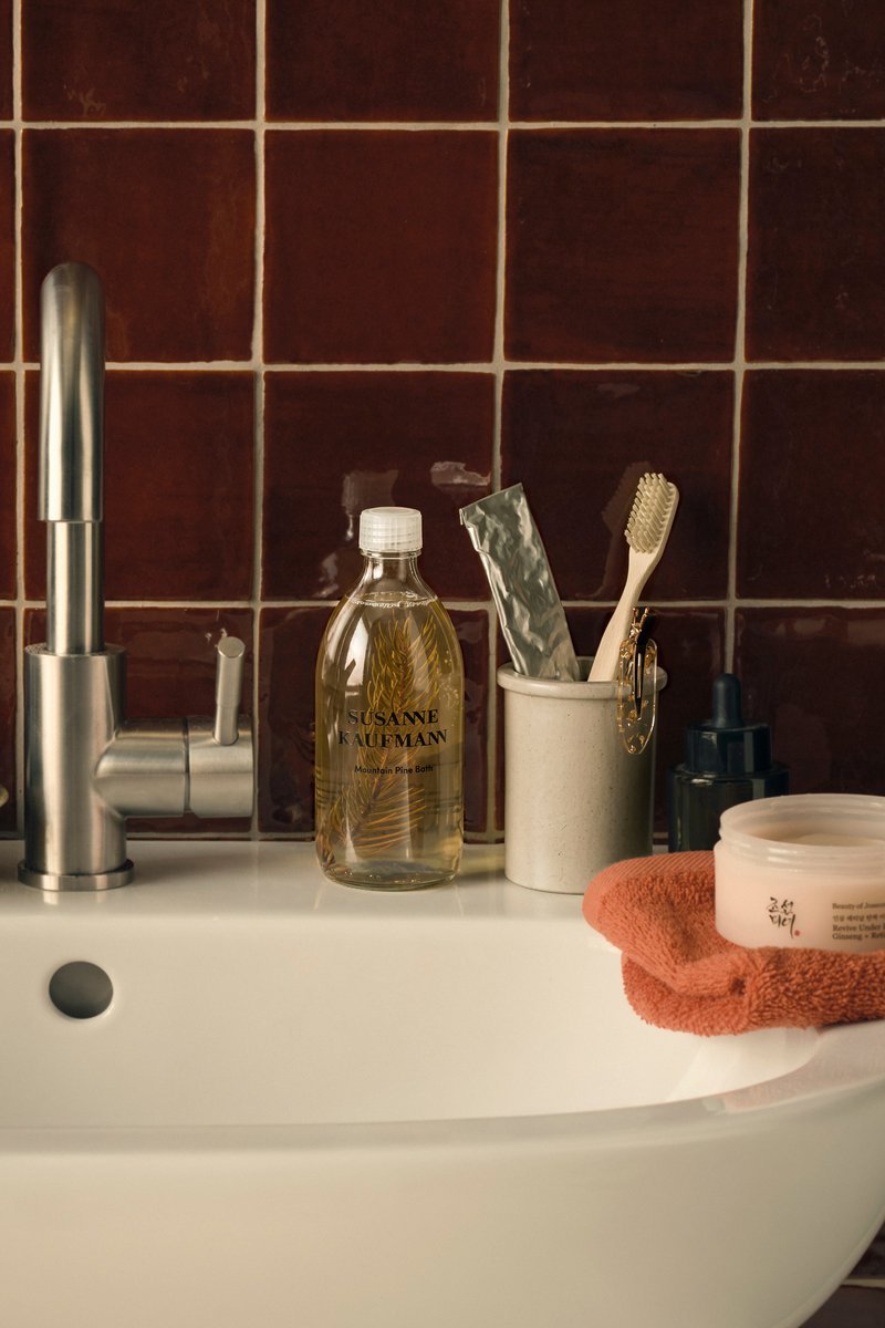 Bathroom sink with a silver faucet, clear bottle labeled Susanne Kaufmann, toothbrush in a holder, dark bottle, and an orange towel with a cream container.