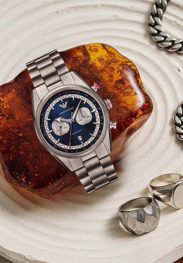 Ρολόι χρονογράφου από ανοξείδωτο χάλυβα με μπλε καντράν, ασημένιους υποκαντράν και κόκκινες λεπτομέρειες, τοποθετημένο σε κεχριμπαρένιο λίθο, δίπλα σε υφασμένα ασημένια δαχτυλίδια.