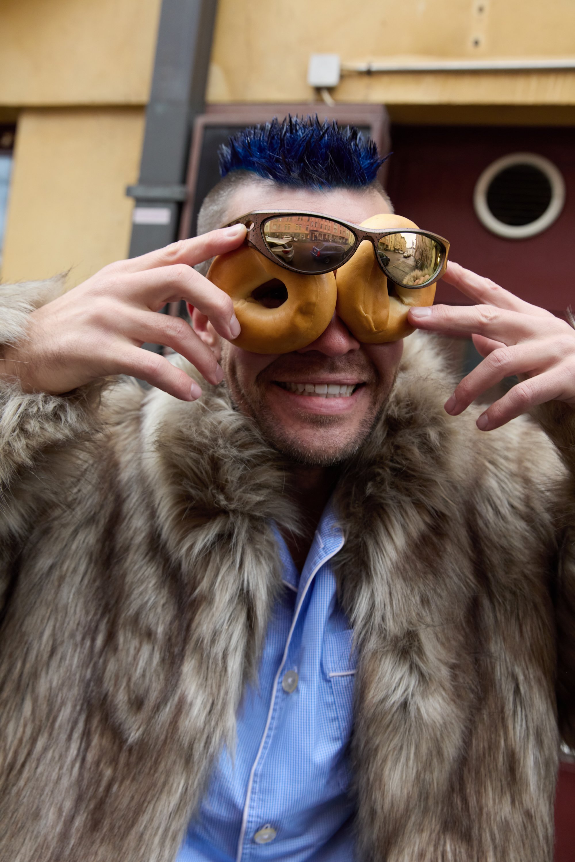 Mies, joka pukeutunut tekoturkistakkiin ja vaaleansiniseen paitaan, silmillään donitsin muotoiset peililasit, poseeraa nostetuin käsin.