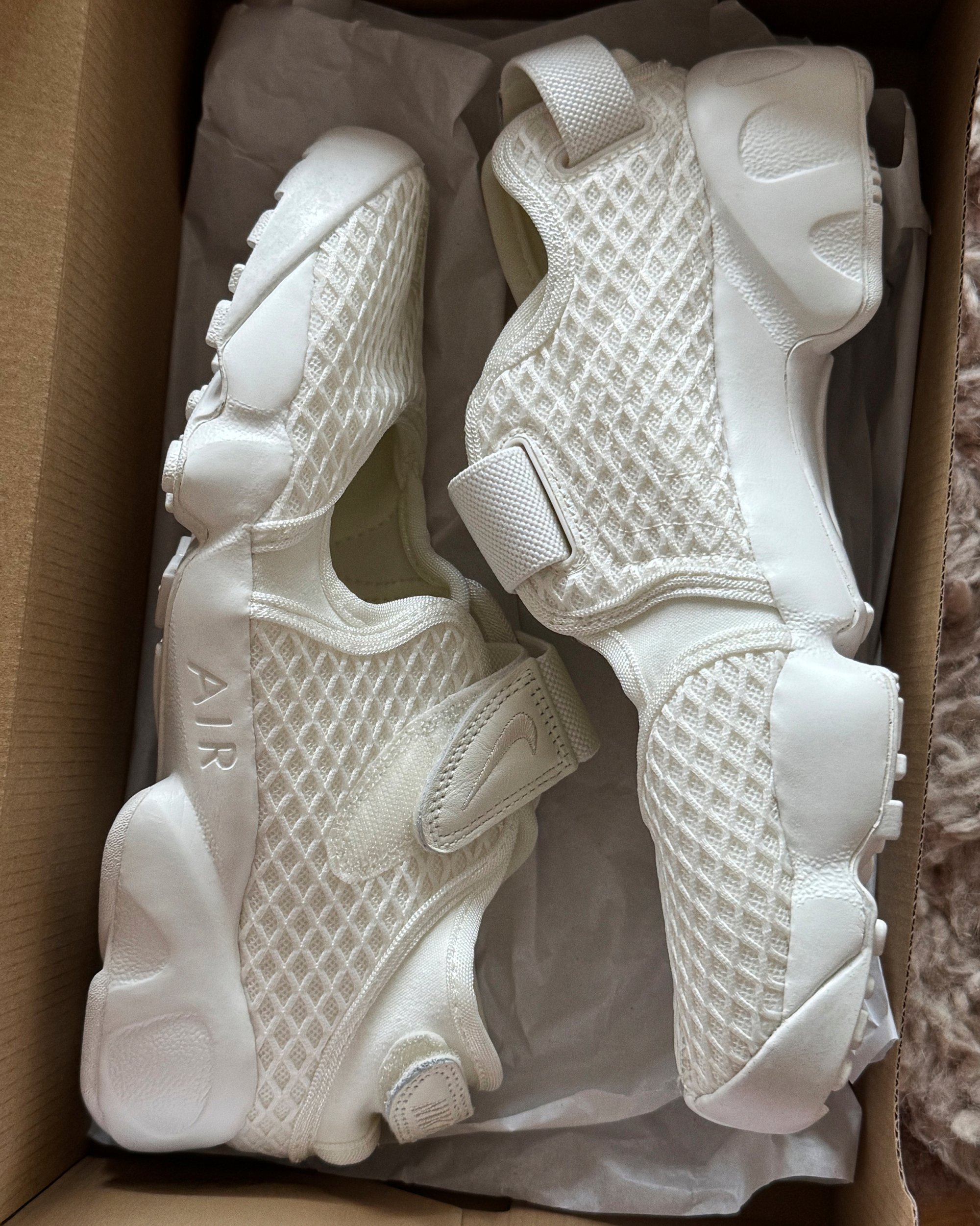 Zapatillas deportivas blancas con un diseño de malla y cinta de velcro, colocadas dentro de una caja de cartón forrada con papel de seda blanco.