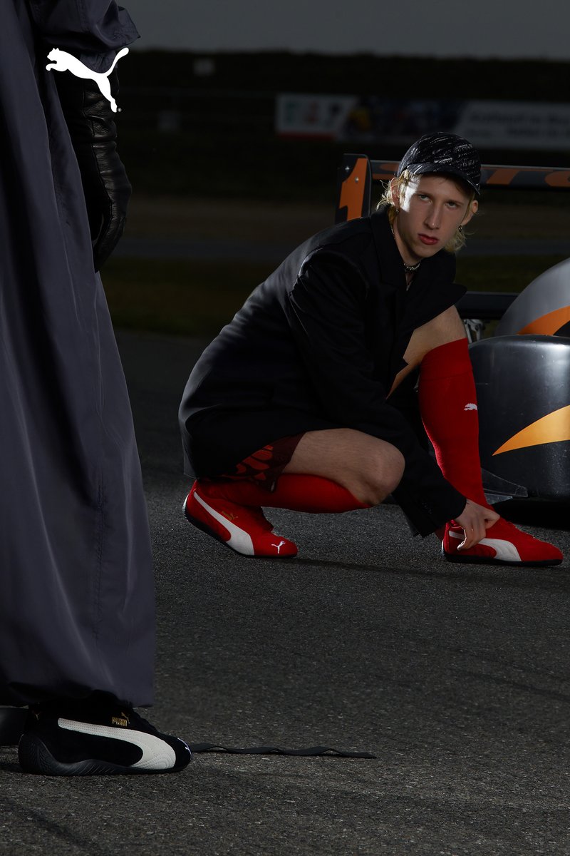 Een persoon knielt naast een raceauto en past hun rode schoeisel aan, terwijl een andere figuur in een donkere outfit in de buurt staat en de situatie observeert.