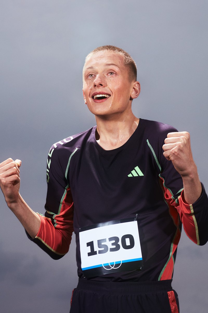 A young male athlete, wearing a black Adidas shirt and bib number 1530, looks upward, clearly exhausted and focused, against a gray background.
