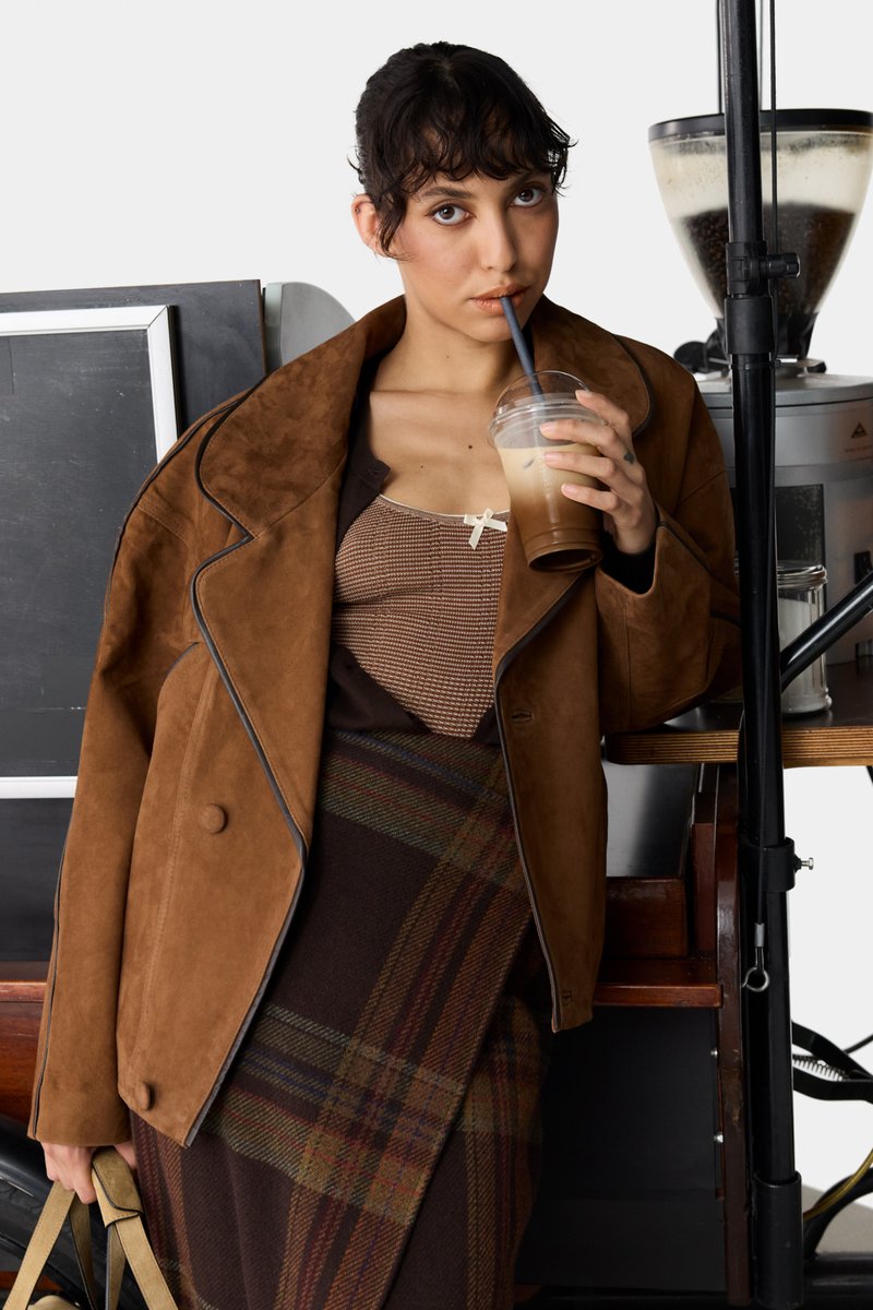 Braune Wildlederjacke im Oversized-Stil mit einem Reverskragen, über einem gemusterten Oberteil und einem karierten Rock getragen. Leichtes Getränk in der Hand, in einem Café sitzend.