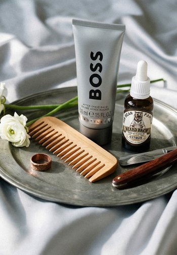 Bálsamo aftershave, aceite para barba, peine de madera, navaja de afeitar y anillo de cobre sobre una bandeja metálica con flores blancas y fondo de tela.