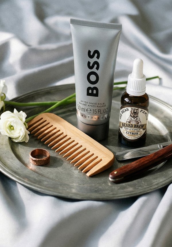 Bálsamo aftershave, aceite para barba, peine de madera, navaja de afeitar y anillo de cobre sobre una bandeja metálica con flores blancas y fondo de tela.