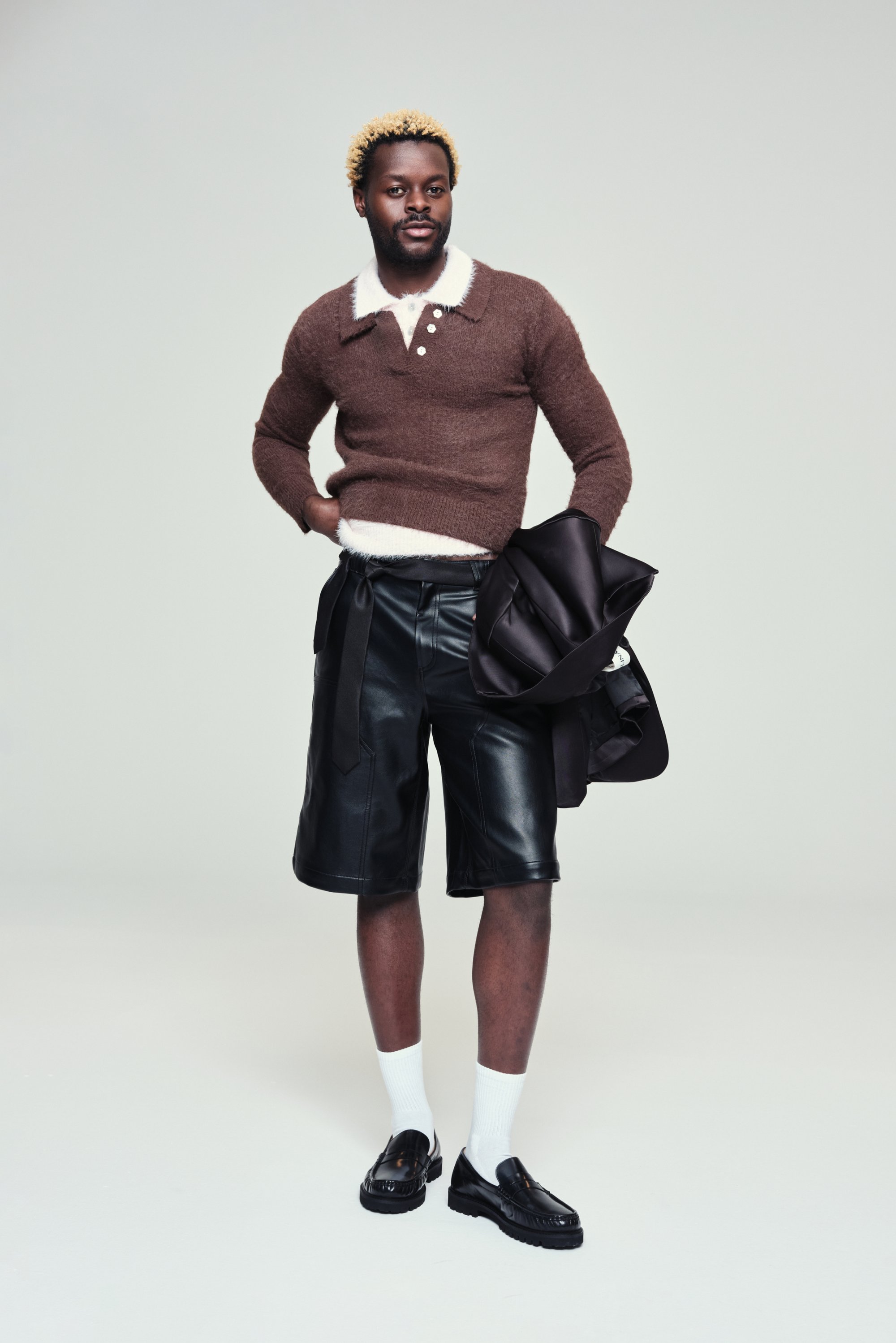 Uomo con capelli neri corti e punte bionde si trova in posa sicura, indossando un maglione marrone e pantaloni corti di pelle nera, tenendo in mano una giacca.
