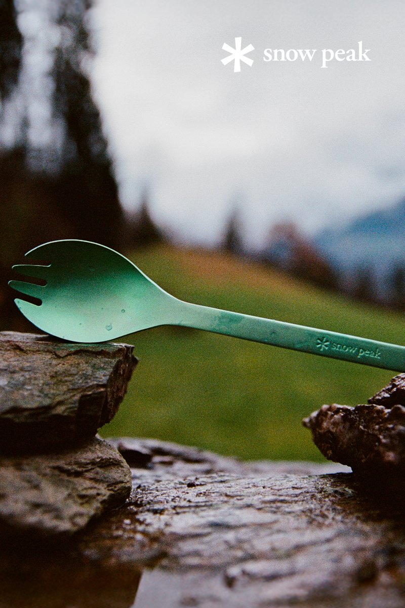 A green spork rests on a rock, with blurred trees and mountains in the background, showcasing the Snow Peak brand logo.