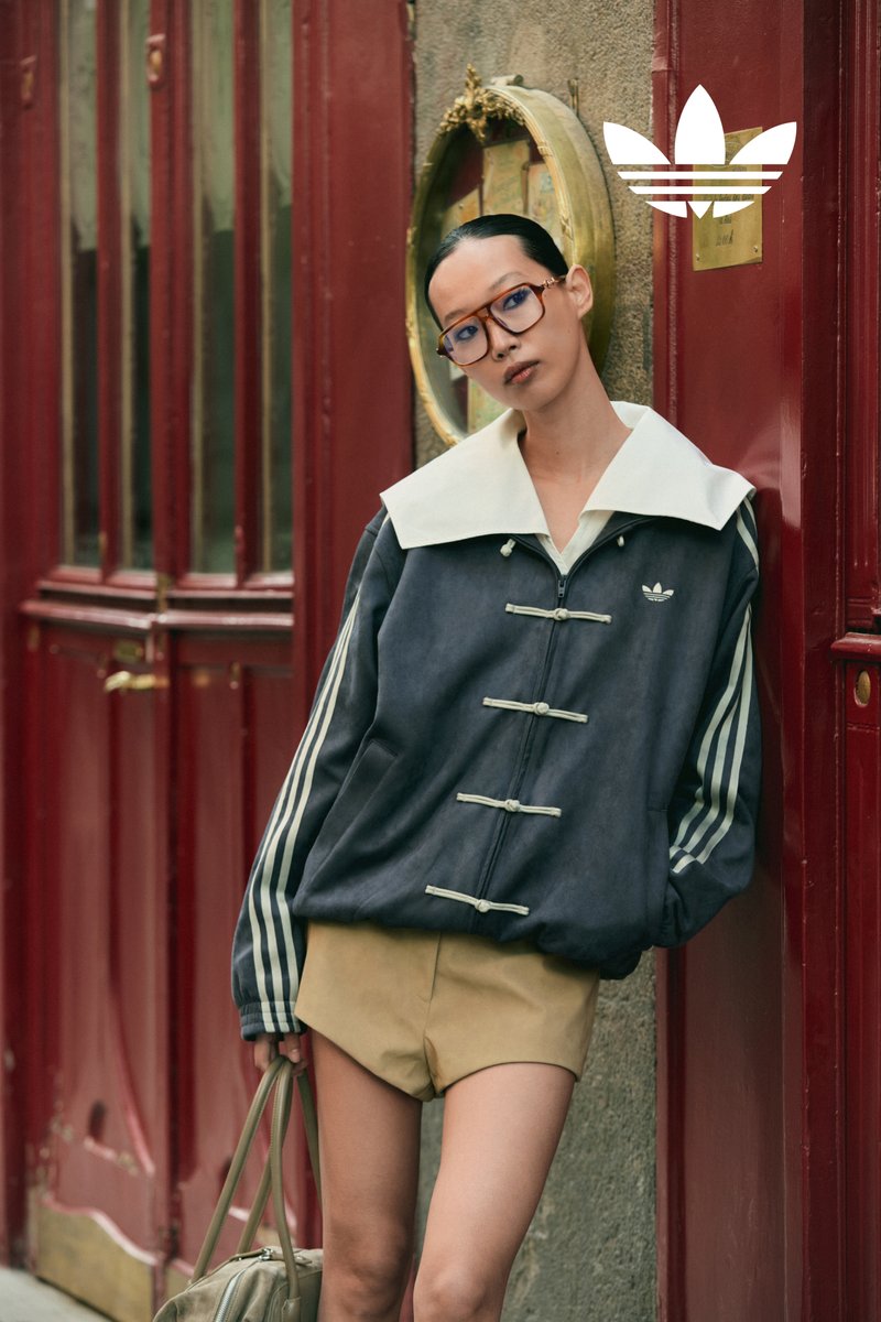 Frau trägt übergroße dunkle Jacke mit weißem Kragen, beige Shorts, Brille und hält eine Tasche, lehnt an einer roten Tür mit rundem Spiegel.