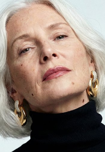 Donna matura con capelli bianchi, che indossa grandi orecchini a cerchio in oro e diamanti e un dolcevita nero, che guarda leggermente verso l'alto con un'espressione neutra.