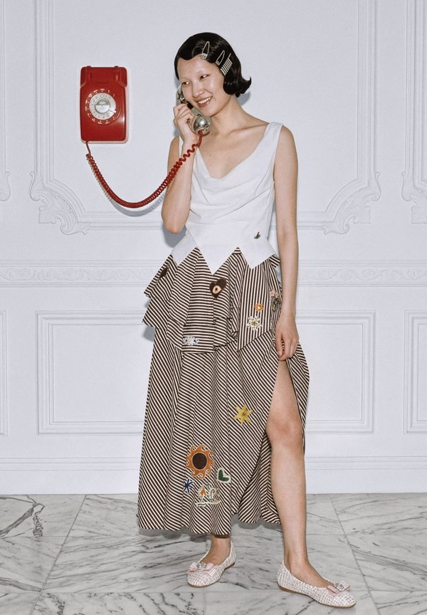 Γυναίκα με τσιμπιδάκια στα μαλλιά χαμογελά κρατώντας το ακουστικό ενός κόκκινου περιστροφικού τηλεφώνου τοίχου, φορώντας λευκή μπλούζα και ριγωτή φούστα με patches.