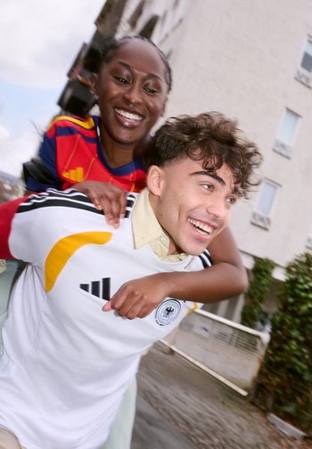 Jonge man in een wit en geel sportshirt geeft een rugzakritje aan een glimlachende vrouw in een rood en blauw sportshirt buiten bij een gebouw.