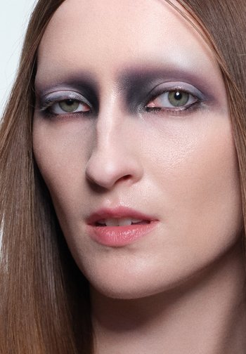 Close-up van een gezicht met een bleke huid, lange bruine haren en dramatische zwarte en grijze oogmake-up met een glanzende, natuurlijke lipkleur.