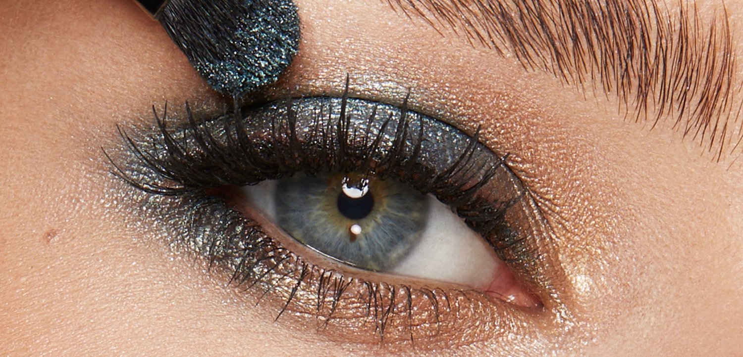 Blaues Auge mit langen Wimpern, das mit einem Make-up-Pinsel auf dem oberen Augenlid mit dunklem metallischem Lidschatten schattiert wird.