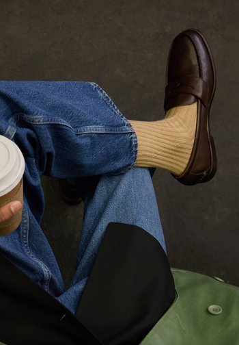 Persoon draagt een blauwe spijkerbroek, beige geribbelde sokken en donkerbruine loafers, houdt een koffiekop vast en zit met gekruiste benen.